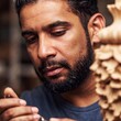 © LIGHTFIELD STUDIOS - A macro shot of a Latino artisan carefully carving a wooden sculpture in his workshop. The extreme close-up captures the intricate details of the carving tools and wood shavings around him.