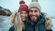 ©  Shomixer - A happy couple with winter hats smiling warmly, standing close against a beach backdrop. The image captures love, joy, and the essence of togetherness.