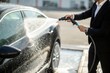 © SerPak - A man dressed in a formal suit is washing a black car with a hose, spraying water over its surface in an open car wash area. The sunlight highlights the shiny surface of the vehicle as he meticulously