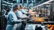 © primopiano - Workers in protective clothing and hairnets in a food processing facility operating machinery with trays of food, focusing on hygiene and quality control in a production line environment.
