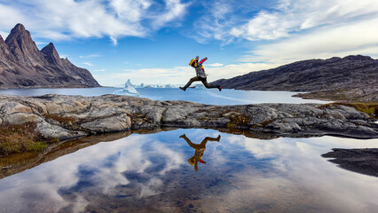 Wall Mural - young man jumping across rocks in bear islands, scoresby sund, greenland. there is a perfect mirror 