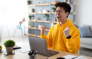 Excited asian man emotionally celebrating success using laptop in home office, sitting at desk raising hands with joy, shaking fists, received good news, got promotion, finished project, free space