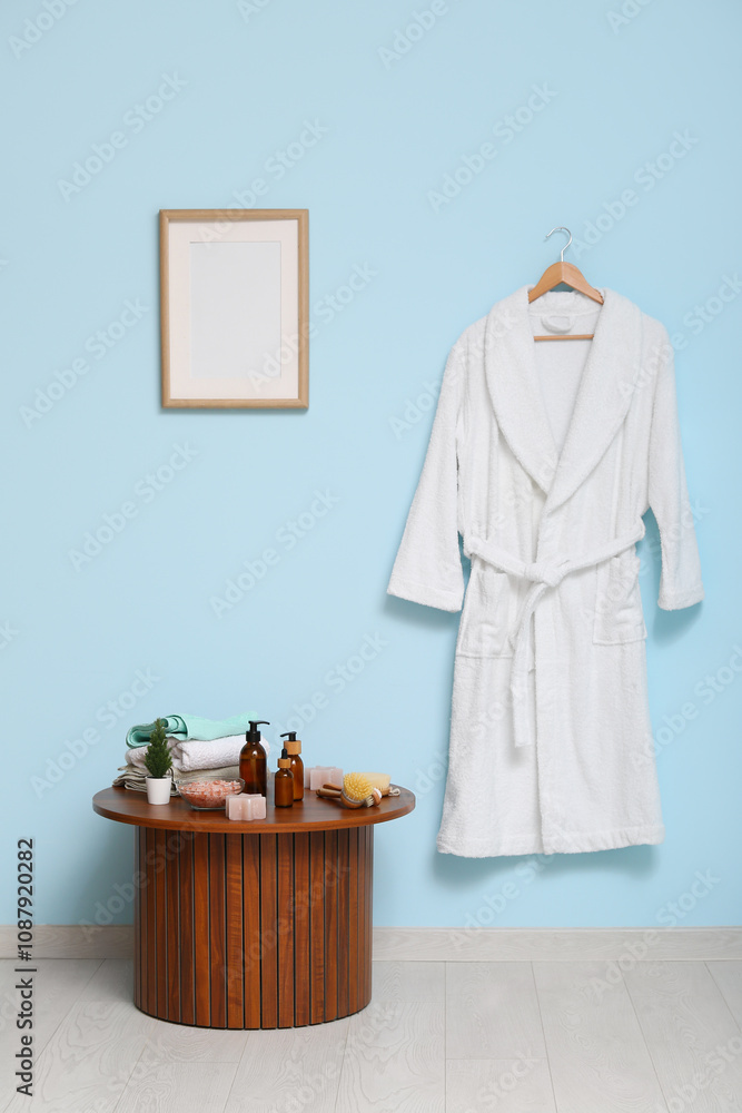 White bathrobe hanging on blue wall and different bath supplies on table in bathroom