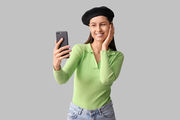 Wall Mural - Beautiful young woman in stylish black beret hat taking selfie on grey background