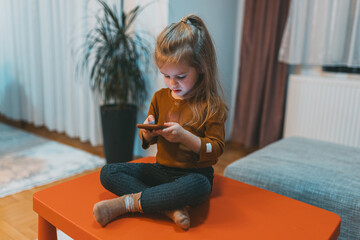 The image depicts a curious girl looking at a smartphone while sitting cross-legged on an orange table, indoors, surrounded by a cozy and warm home environment with decor.