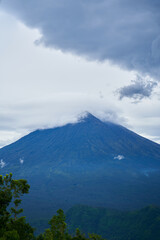 Wall Mural - Panorama of Mount Agung and rice fields on the island of Bali. View of the mountain against a background of palm trees and a cornfield. Panorama of Agung volcano covered with clouds on a rainy day.