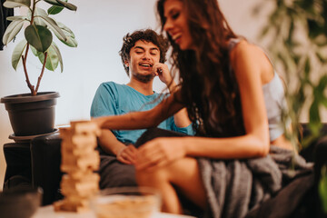 Poster - A young man and woman share a laugh while playing a block tower game at home. The warm setting hints at friendship and joy in a relaxed environment.