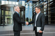 © Jose Calsina - experienced senior executive warmly greeting younger business partner with confident handshake outside modern corporate building entrance, symbolizing successful mentorship, professional collaboration