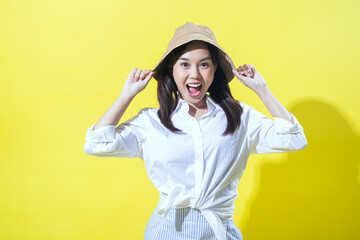 Asian woman wearing a white shirt and striped skirt, playfully holding a beige bucket hat with a cheerful expression against a vibrant yellow background, radiating joy and positivity.