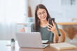 © oatawa - Smiling young asian woman using laptop while holding a cup of coffee, talking to company client or colleague on mobile smartphone at home. lifestyle