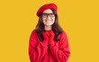 © Studio Romantic - Portrait of beautiful happy smiling brunette young woman. Excited girl wearing eyeglasses, red beret, gloves and sweatshirt holding hands on chest feeling joy and delight on yellow background