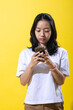 © Daniel - An Asian young woman with black hair wearing a white t-shirt and brown pants, holding a smartphone with both hands. She is focused on the phone, against a vibrant yellow background