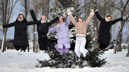 Five joyful girls are gleefully leaping together in a snowy park beside a pine tree in the winter season