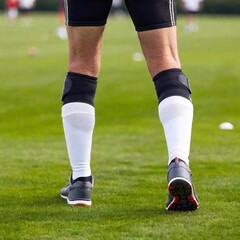 Wall Mural - Elderly player leg, highlighting knee brace in sharp focus, with the grassy field and other players in the distance