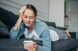 © Ilona - A beautiful woman lying on the sofa at home scrolls news on social networks on her phone.