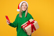 © ViDi Studio - Merry young woman wear green sweater Santa hat hold present box with gift use mobile cell phone look aside isolated on plain yellow background. Happy New Year, celebration Christmas holiday concept.