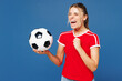 © ViDi Studio - Young fun woman fan wear basic red t-shirt cheer up support football sport team hold in hand soccer ball watch tv live stream do winner gesture celebrate clench fist isolated on plain blue background