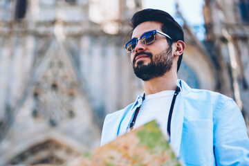 Wall Mural - Bearded tourist in sunglasses looking straight standing in downtown with old construction on background.Traveler holding map for searching showplaces while strolling on streets of architectural city