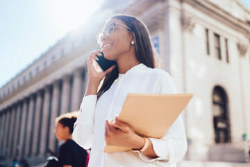 Successful african american student of faculty of law strolling in urban setting communicating with friends on smartphone.Positive female office worker with folder calling on telephone in downtown