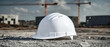 © Anoo - White construction hard hat rests on gravel, surrounded by concrete slabs and blurred cranes under soft gray lighting in an architectural site