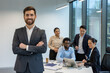 © Liubomir - Confident businessman standing in front of diverse team engaged in office meeting. Group includes professionals discussing at table with laptop and smartphone. Bright, modern workspace.