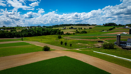 Wall Mural - Green fields and brown patches extend across the countryside, framed by trees and a distant hilly backdrop. Bright clouds float above, casting shadows on the vibrant landscape during daytime.