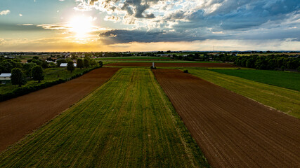 Wall Mural - A vibrant countryside vista showcases vast fields of varying shades of green and brown under a dramatic sky. The sun sets, casting a warm glow on the agricultural land and peaceful surroundings.