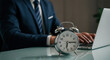 © TAMER YILMAZ - Close-up alarm clock on desk, businessman in suit using laptop in blurred area, working with correspondence in background, entrepreneur involved in work process, workday, time management concept.