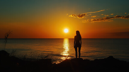 Wall Mural - A woman stands on a rock overlooking the ocean at sunset