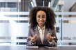 © Tetiana - Portrait of a smiling young African-American woman sitting in the office at a desk in a headset, talking and gesturing with her hands to the camera