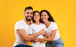 © Prostock-studio - Arab Family. Happy Middle-Eastern Parents Hugging Their Kid Daughter Smiling To Camera On Yellow Background. Studio Shot Of Spouses Embracing Little Girl Expressing Their Love To Child