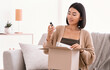 © Prostock-studio - Beauty Box Concept. Portrait of smiling asian woman received delivery package, holding foundation base for makeup, unpacking cardboard parcel with cosmetics product, sitting on the couch at home