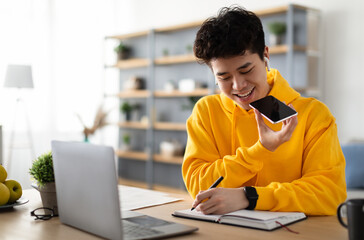 Work From Home. Young Asian Guy Talking On Mobile Phone And Taking Notes, Using Voice Assistant, Writing In Notebook. Smiling Man Sitting At Desk With Laptop At Home Office, Wearing Earbuds