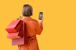 © Pixel-Shot - Young woman in autumn clothes with mobile phone and shopping bags on yellow background, back view