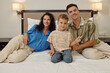 © DragonImages - Happy family smiling and relaxing on bed with modern wall lamps behind them creating cozy ambience in bedroom. Young boy sitting between smiling parents