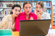 © Raquel - Three women in a library react with surprise while viewing a laptop. The scene conveys emotions of amazement, curiosity, or shock, surrounded by bookshelves and study materials.
