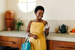 © (JLco) Julia Amaral - Cheerful woman smiling and holding a vegetable bag while using her mobile phone in the kitchen