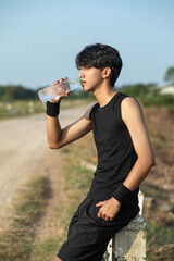 Asian Young man in black sportswear, tired, drinking water after jogging, outdoors