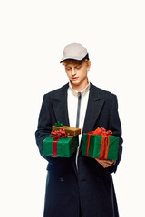 Dressed in trendy winter attire, a young man stands holding two beautifully wrapped Christmas gifts. His fashionable coat and cap reflect the festive spirit.