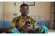 © N Joy Art  - African male mental health professional in colorful traditional wear, holding a smartphone in a relaxed indoor environment, demonstrating app-based mental health support for personal well-being.