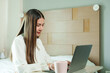 © Ekkasit A Siam - Asian young woman sitting in bedroom with modern wooden decor, using laptop, wearing white blouse, showing serious expression, warm lighting creating comfortable work-from-home setup.