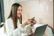 © Ekkasit A Siam - Asian woman smiling while holding credit card and using laptop in cozy bedroom, white blouse, light wooden decor, representing modern online shopping and digital transaction concept.