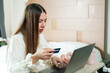 © Ekkasit A Siam - Asian young woman holding credit card, appearing surprised while shopping online with laptop in bedroom, white blouse, light decor, highlighting digital finance and e-commerce.