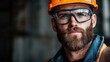 ©  Shomixer - A focused worker with a beard is wearing protective goggles and an orange hard hat while standing in an industrial environment, emphasizing safety and professionalism.