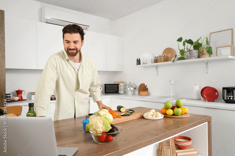 Handsome bearded man with laptop cooking in kitchen