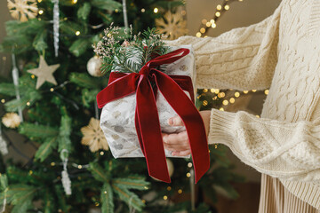 Wall Mural - Merry Christmas and Happy Holidays. Hands in sweater holding modern christmas gift box with red ribbon and fir branch on background of stylish christmas tree with lights.