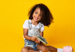 © Prostock-studio - Cute afro girl holding bunch of money, sitting over yellow background