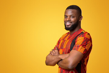 Wall Mural - Confident smiling african american guy in bright traditional t-shirt over yellow studio background, copy space