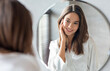 © Prostock-studio - Self-Care Concept. Young Attractive Woman Looking At Mirror In Bathroom, Pretty Female Touching Face And Smiling To Reflection, Enjoying Her Beautiful Smooth Skin, Selective Focus, Closeup