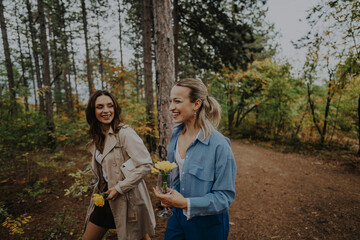 Poster - Two friends are happily strolling through a vibrant autumn forest, sharing laughter and camaraderie. They are holding flowers and dressed for a cool day, surrounded by colorful trees.